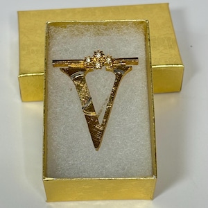 Peut inclure: Broche en forme de lettre V dorée avec des strass clairs en forme d'étoile. La broche est dans une petite boîte dorée.