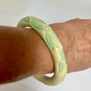 May include: Cream-colored bangle bracelet with carved floral and leaf design, accented with light blue. The bracelet is a thick, rounded shape, and the floral pattern is repeated around the entire circumference.