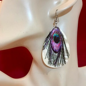 Puede incluir: Un par de pendientes colgantes con una base blanca en forma de concha y un diseño de pluma de pavo real colorido.