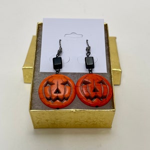 May include: A pair of orange and black pumpkin earrings in a gold gift box. The earrings are made of carved stone and have a black bead at the top.