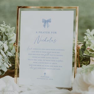 May include: A framed print with the text "A Prayer for Nicholas" in elegant script. The print features a blue bow illustration and a prayer, with the word "Baptized" and the date November 13, 2023 at the bottom. The frame is gold.