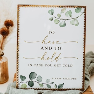 May include: A rectangular glass sign with a metallic gold frame featuring green watercolor eucalyptus leaves and the text: TO have AND TO hold IN CASE YOU GET COLD, PLEASE TAKE ONE. The sign is displayed in a vertical orientation.
