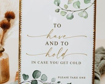 May include: A rectangular glass sign with a metallic gold frame featuring green watercolor eucalyptus leaves and the text: TO have AND TO hold IN CASE YOU GET COLD, PLEASE TAKE ONE. The sign is displayed in a vertical orientation.