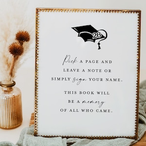 May include: A graduation guest book sign with a gold frame and white background. The sign features a black graduation cap graphic with the number 26. Text on the sign reads: "Pick a page and leave a note or simply sign your name. This book will be a memory of all who came."