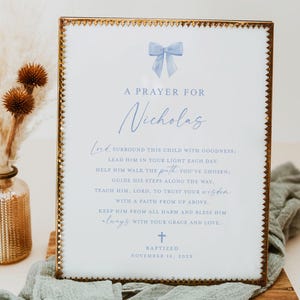 May include: A framed print with a gold border and a light blue bow illustration at the top. The text reads "A Prayer for Nicholas" and includes a prayer. The print is on a wooden board with a sage green cloth.