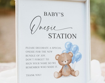 May include: A framed sign with the text "BABY'S Onesie STATION" and a teddy bear illustration holding blue balloons. The sign includes instructions to decorate a onesie for the new baby and sign it. The sign is on a white cloth.