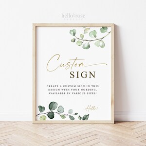 May include: A white sign with a gold border and green eucalyptus leaves. The sign says "Custom Sign" and "Create a custom sign in this design with your wording. Available in various sizes!" with the text "Hello!" at the bottom.