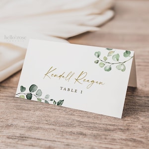 May include: A white place card with the text "Kendall Reagan" in gold script, and "TABLE 1" in a smaller font. The card is decorated with watercolor eucalyptus leaves. The "hello rose" logo is in the upper left corner.