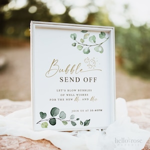 May include: A white sign with a gold frame and green eucalyptus leaves. The sign says "Bubble Send Off" and "Let's blow bubbles of well wishes for the new Mr. and Mrs. Join us at 10:40PM".