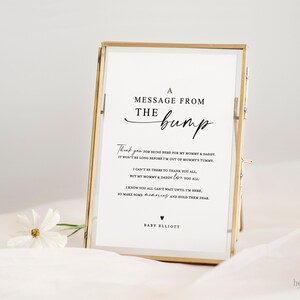 Op de afbeelding: Een wit ingelijst bord met een boodschap van de bult. Het bord luidt: "Een boodschap van de bult. Bedankt dat je er bent voor mijn mama en papa. Het duurt niet lang meer voordat ik uit mama's buik ben. Ik kan er niet zijn om jullie allemaal te bedanken, maar mijn mama en papa houden van jullie allemaal. Ik weet dat jullie allemaal niet kunnen wachten tot ik er ben, dus maak wat herinneringen en bewaar ze goed. Baby Elliott."