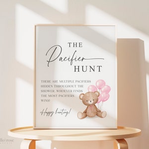 May include: A white sign with the text "THE Pacifier HUNT" in black script font. There are multiple pacifiers hidden throughout the shower. Whoever finds the most pacifiers wins! Happy hunting!"  A brown teddy bear with pink bows is holding pink balloons.