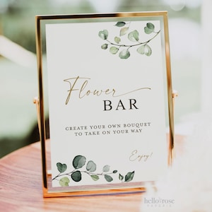 May include: A gold framed sign with a white background and green watercolor leaves. The sign reads "Flower Bar" and "Create your own bouquet to take on your way". The sign also says "Enjoy!" and has the text "hello rose paperie" at the bottom.