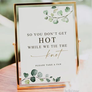 May include: A gold-framed sign with a white background and green watercolor eucalyptus leaves. The sign reads "So you don't get hot while we tie the knot. Please take a fan."
