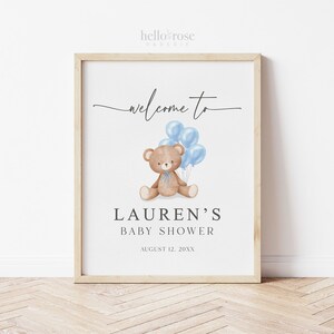 May include: A framed baby shower sign with a light wood frame. The sign features the text "welcome to" in a script font, with a watercolor illustration of a teddy bear holding blue balloons. The text "LAUREN'S BABY SHOWER" is below the illustration.