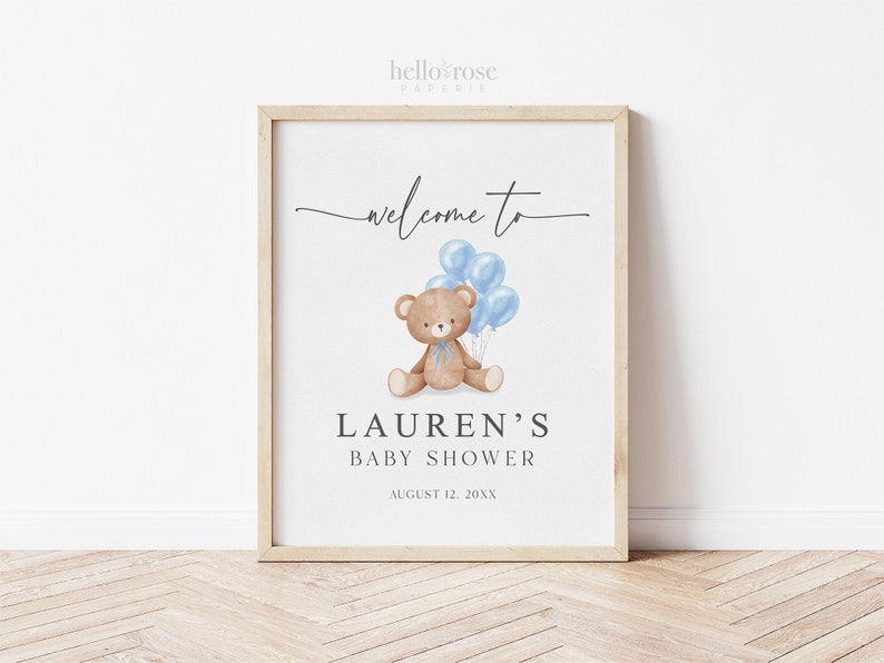 May include: A framed baby shower sign with a light wood frame. The sign features the text "welcome to" in a script font, with a watercolor illustration of a teddy bear holding blue balloons. The text "LAUREN'S BABY SHOWER" is below the illustration.