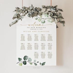 May include: A wedding seating chart with a eucalyptus and greenery design. The chart is printed on white cardstock and features the text "Welcome to our wedding, February 12, 2023, Micah and Caroline".