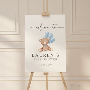 May include: A baby shower welcome sign on an easel. The sign features a watercolor illustration of a teddy bear holding blue balloons. The text reads "welcome to" above the illustration, and "LAUREN'S BABY SHOWER" below, with the date.