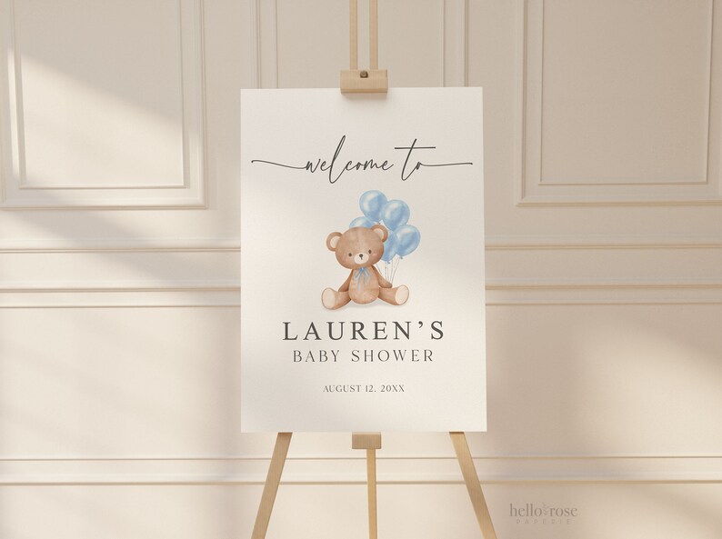 May include: A baby shower welcome sign on an easel. The sign features a watercolor illustration of a teddy bear holding blue balloons. The text reads "welcome to" above the illustration, and "LAUREN'S BABY SHOWER" below, with the date.