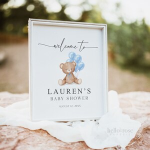 May include: A framed baby shower sign with the text "welcome to" at the top. Below is a brown teddy bear holding blue balloons. The sign reads "Lauren's Baby Shower" and the date "August 12, 20XX".