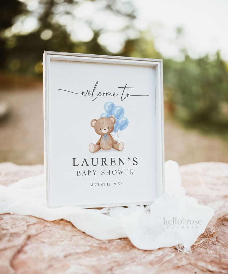 May include: A framed baby shower sign with the text "welcome to" at the top. Below is a brown teddy bear holding blue balloons. The sign reads "Lauren's Baby Shower" and the date "August 12, 20XX".