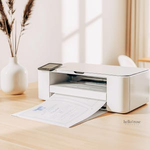 May include: A white printer on a light wood surface, printing a baby shower invitation. The invitation has a blue bow design and the text "Baby Shower". A small black device sits on top of the printer. A white vase with dried plants is to the left.