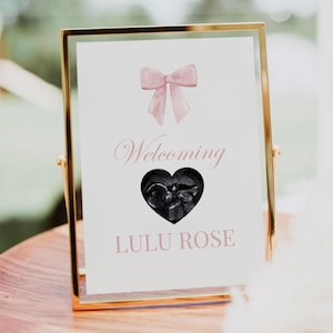 May include: A white sign with a pink bow at the top and a black and white ultrasound image in a heart shape. The sign says "Welcoming Lulu Rose".