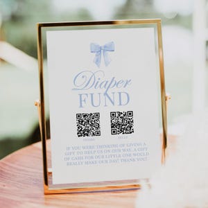 May include: A white sign with a blue bow and the text "Diaper Fund" with two QR codes labeled "Venmo" and "Zelle". The sign reads "If you were thinking of giving a gift to help us on our way, a gift of cash for our little one would really make our day! Thank you!"