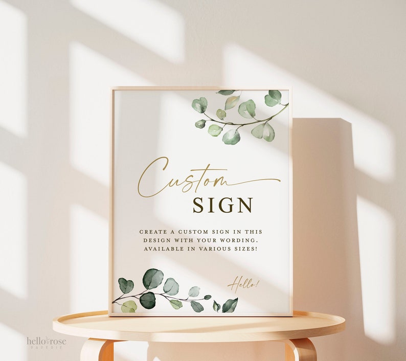 May include: A white sign with a gold frame and green eucalyptus leaves. The sign says "Custom Sign" and "Create a custom sign in this design with your wording. Available in various sizes!" with the word "Hello!" at the bottom.