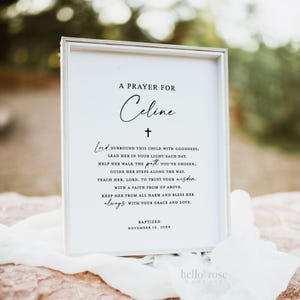 May include: A white framed print with a black cross and the text "A Prayer for Celine" and a prayer for the child. The text reads "Lord, surround this child with goodness; lead her in your light each day. Help her walk the path you've chosen; guide her steps along the way. Teach her, Lord, to trust your wisdom. With a faith from up above. Keep her from all harm and bless her always with your grace and love." The print also includes the text "Baptized November 12, 20XX".