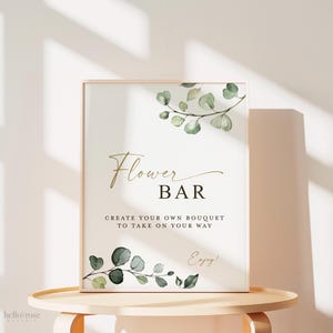Pode incluir: Uma placa retangular numa moldura de madeira clara com ilustra&ccedil;&otilde;es de folhas de eucalipto verdes no topo e na base. O texto diz Flower BAR, CREATE YOUR OWN BOUQUET TO TAKE ON YOUR WAY e Enjoy! em letras douradas e pretas.