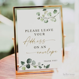 May include: A white sign with gold lettering and green watercolor leaves. The sign reads "Please leave your address on an envelope. Thank you!"