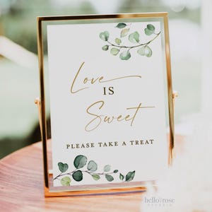 May include: A gold-framed sign with a white background and green watercolor leaves. The sign reads "Love is Sweet" and "Please take a treat".