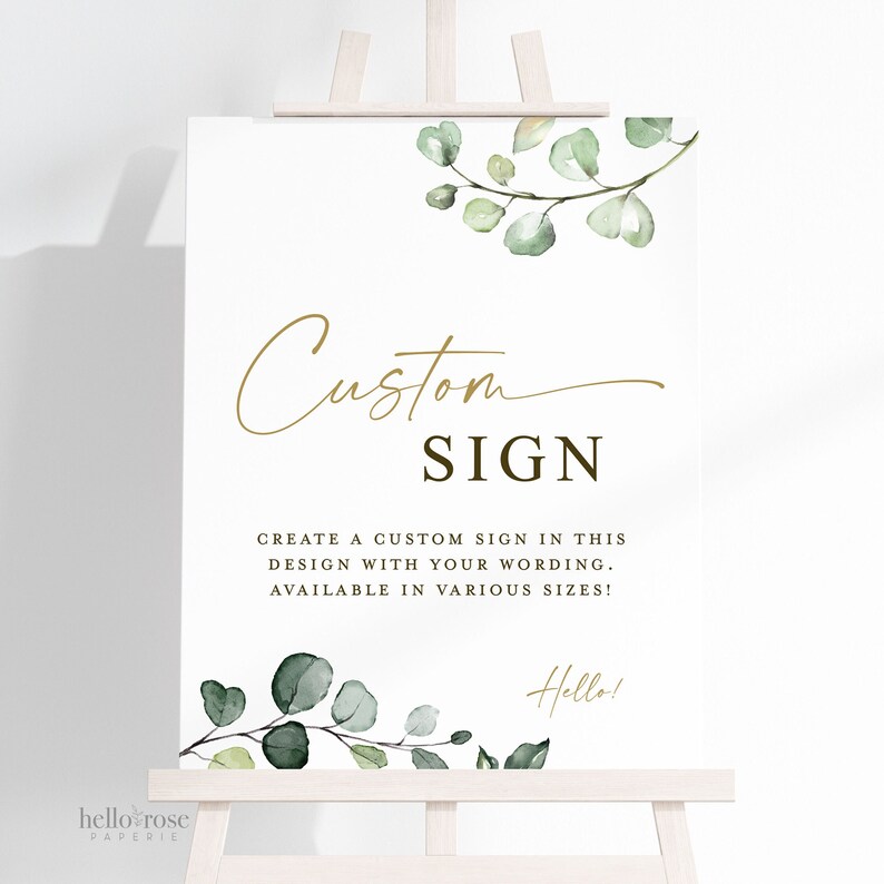 May include: A white sign with a gold script font that says "Custom Sign". The sign also includes the text "Create a custom sign in this design with your wording. Available in various sizes!" and a small green eucalyptus branch with the word "Hello!" in gold script font.