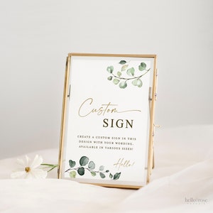 May include: A gold-framed sign with a white background and green watercolor eucalyptus leaves. The sign says "Custom Sign" and "Create a custom sign in this design with your wording. Available in various sizes!" with the text "Hello!" at the bottom.