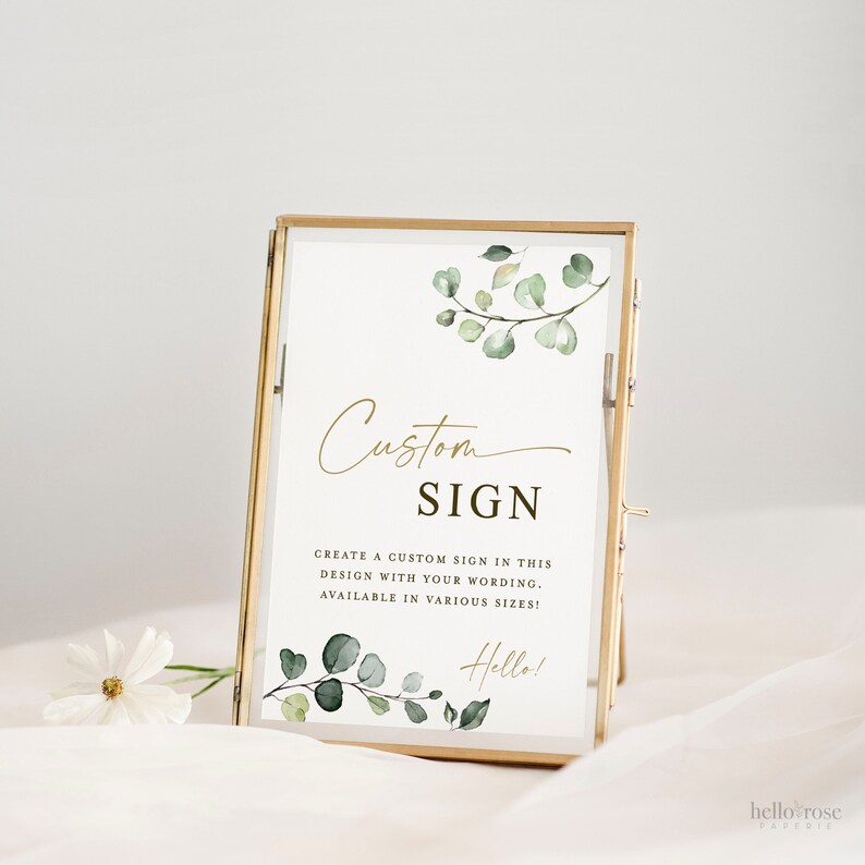 May include: A gold-framed sign with a white background and green watercolor eucalyptus leaves. The sign says "Custom Sign" and "Create a custom sign in this design with your wording. Available in various sizes!" with the text "Hello!" at the bottom.