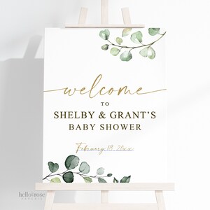 Puede incluir: Un cartel de bienvenida blanco con letras doradas y hojas verdes de acuarela. El cartel dice "Bienvenido a Shelby & Grant's Baby Shower, 18 de febrero de 20xx".