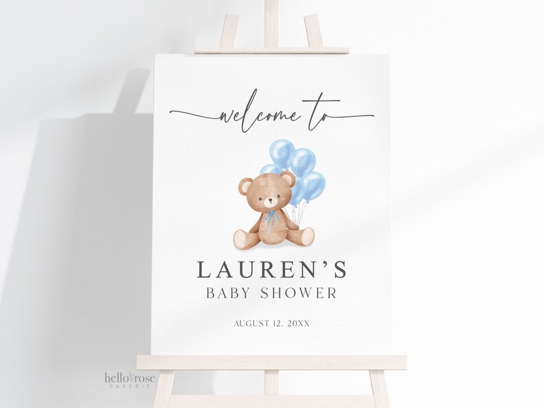 May include: A baby shower sign on an easel. The sign reads "welcome to" in script above a watercolor illustration of a teddy bear holding blue balloons. Below the illustration is "LAUREN'S BABY SHOWER" and the date.