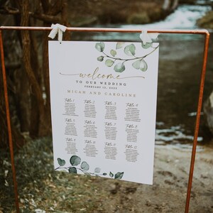May include: A white wedding seating chart with a leafy green watercolor design. The chart lists the names of guests assigned to each table number. The text reads "Welcome to our wedding, February 12, 2025, Micah and Caroline."