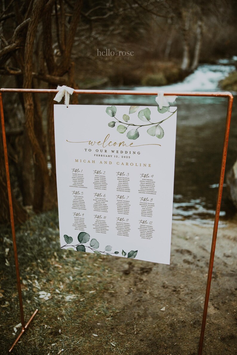 May include: A white wedding seating chart with a leafy green watercolor design. The chart lists the names of guests assigned to each table number. The text reads "Welcome to our wedding, February 12, 2025, Micah and Caroline."