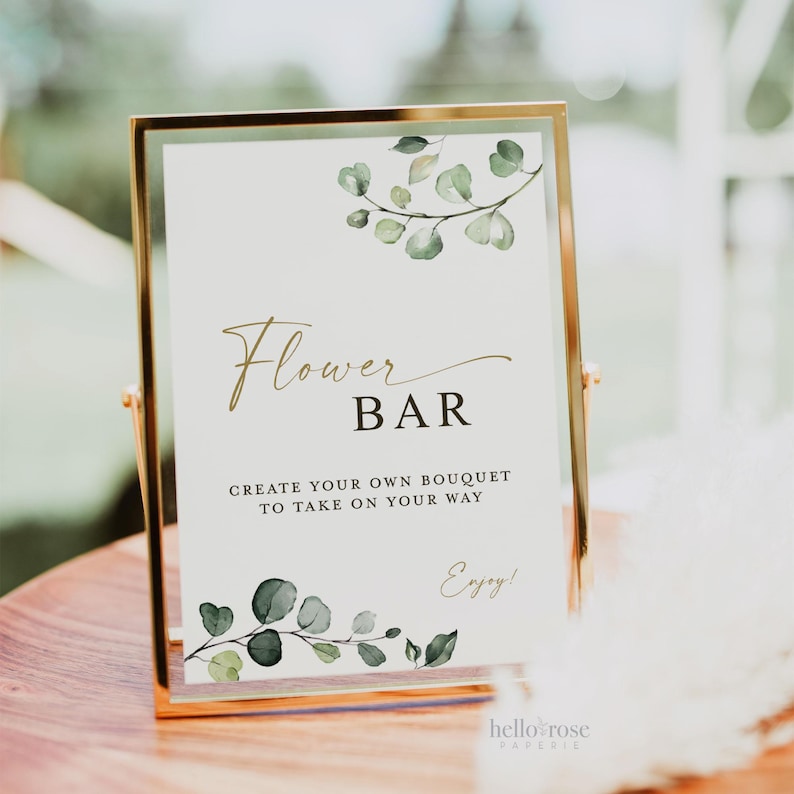 Pode incluir: Uma placa retangular vertical numa moldura de metal dourado fino com o texto Flower BAR CREATE YOUR OWN BOUQUET TO TAKE ON YOUR WAY e Enjoy! decorada com ilustra&ccedil;&otilde;es de folhas de eucalipto verdes nos cantos superiores e inferiores.