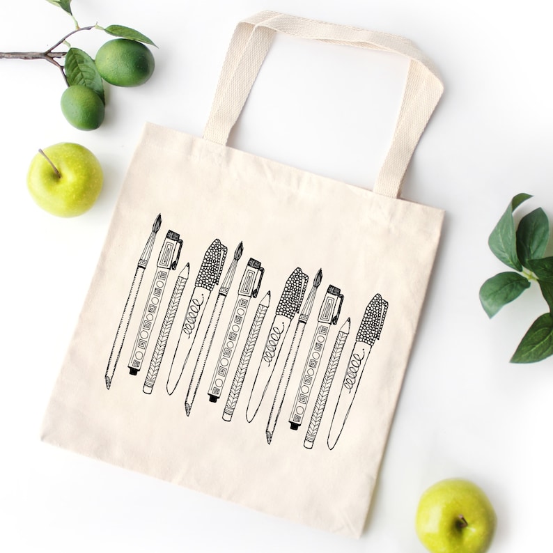 K&ouml;nnte beinhalten: Eine wei&szlig;e Canvas-Tragetasche mit schwarz-wei&szlig;en Illustrationen verschiedener Kunstmaterialien, darunter Pinsel, Stifte und Bleistifte. Die Tasche hat einen schwarzen Griff.