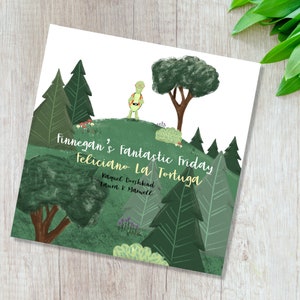 Op de afbeelding: Kinderboekcover met een groene schildpad met bril en rugzak op een groene heuvel. De titel luidt "Finnegan's Fantastic Friday Feliciano La Tortuga". De illustratie bevat bomen en een groen landschap.