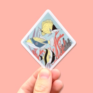May include: A sticker featuring a colorful illustration of three fish swimming in a coral reef. The fish are yellow, blue, and black with white stripes. The coral is red and pink.