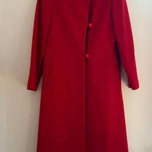 May include: A vibrant red coat with a high collar and three matching buttons. The coat features long sleeves and two front pockets. The coat is hanging on a hanger against a white wall.
