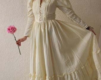 May include: A long, cream-colored dress with a high collar and long sleeves. The dress features a fitted bodice with decorative buttons and a full skirt with multiple ruffled tiers. A pink rose is held in the hand.