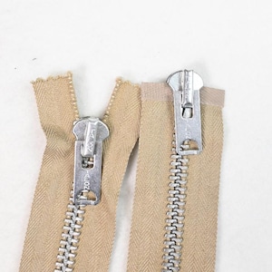 May include: Two beige zippers with silver metal pulls. The zippers are open, showing the teeth and the fabric tape. The zipper pulls are marked with the word "TALON".