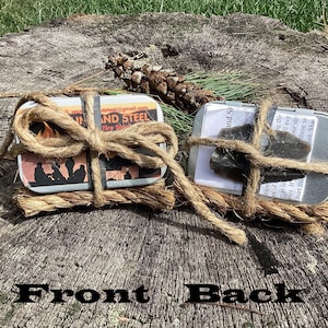 May include: Two rectangular metal tins tied with thick jute twine. The left tin features a label with a fire graphic and the text Flint and Steel. The right tin displays a dark grey stone shard and a white paper label with printed text.