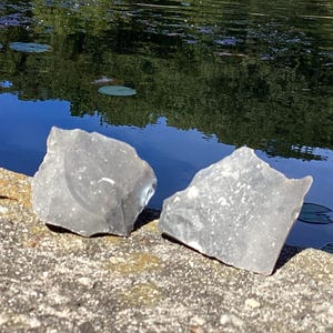 Puede incluir: Dos rocas de pedernal grises de forma irregular descansan sobre una superficie de piedra. Las rocas tienen una textura rugosa y están situadas cerca de una masa de agua con reflejos de árboles y nenúfares.