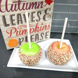 May include: Two caramel apples on sticks, one green and one red, covered in chopped nuts. The apples are on a white plate with a sign that says "Autumn Leaves & Pumpkins Please".