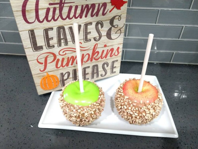 May include: Two caramel apples on sticks, one green and one red, covered in chopped nuts. The apples are on a white plate with a sign that says "Autumn Leaves & Pumpkins Please".